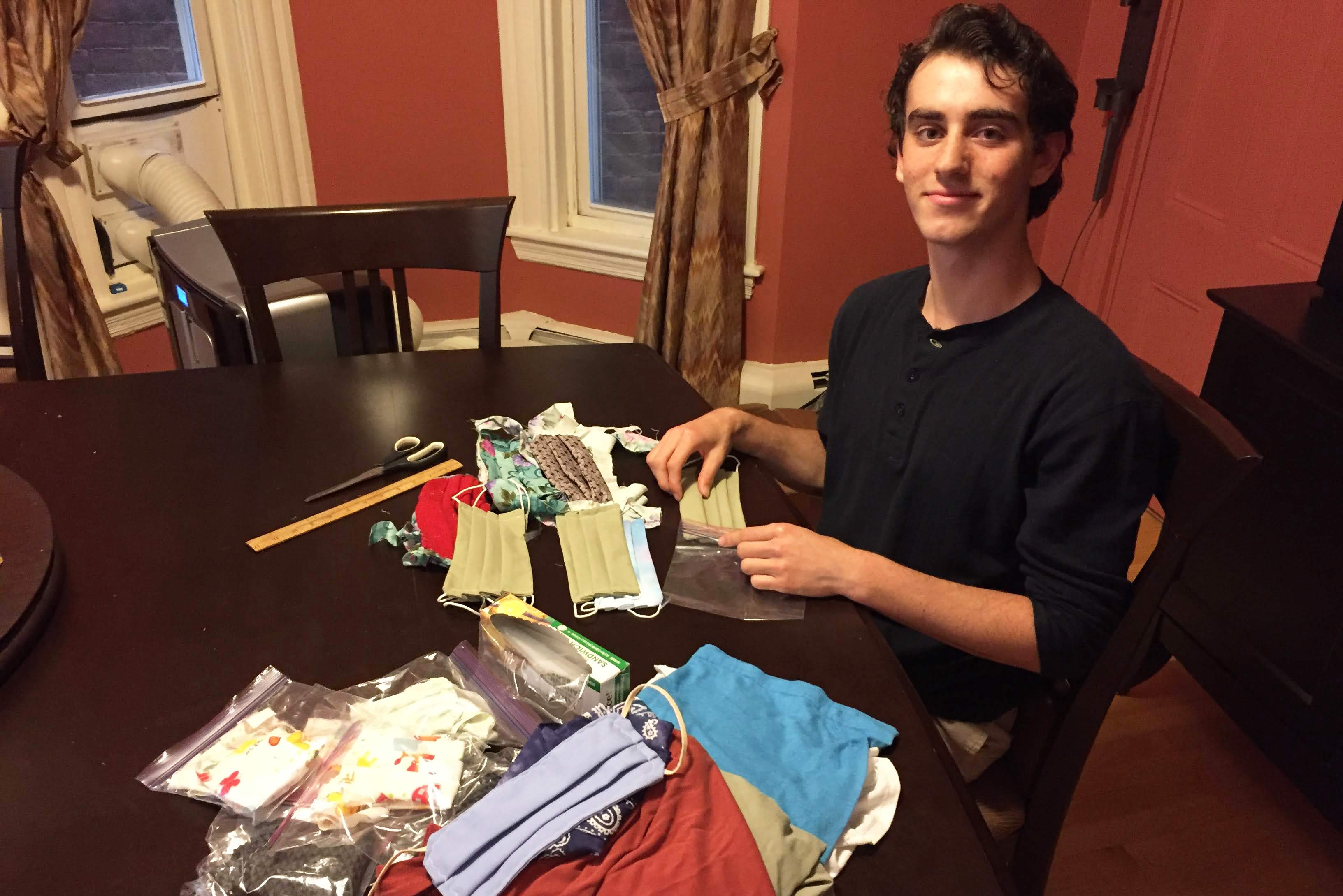 Photo of incoming student Adam Katz (CAS’24), sitting and smiling, as he prepares homemade masks for donation. A pile of masks sit on a kitchen table before him.