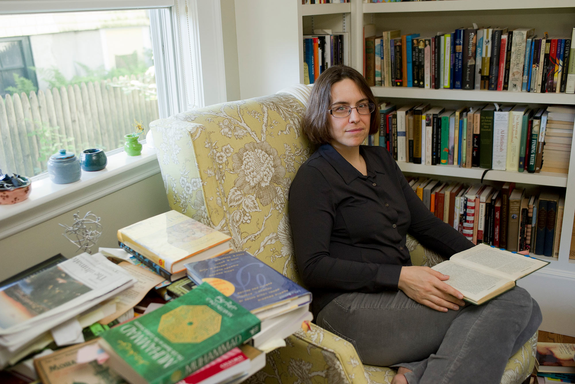 Portrait of CAS Religion Professor Kecia Ali in an office surrounded by books. She shits in an armchair holding a book, and doesn't wear shows.