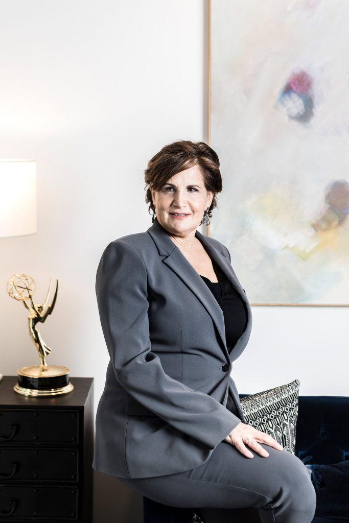 Photo of Brooke Karzen (CFA’84) sitting on a desk in a gray paint suit with a golden globe in the background.