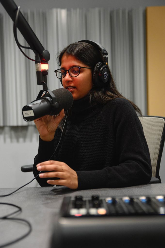 Photo of Sarika Ram (CAS’21) in a black shirt and pants in a podcast recording studio, presumably recording her podcast Criminalized. Sarika touches the mic as she speaks into it.