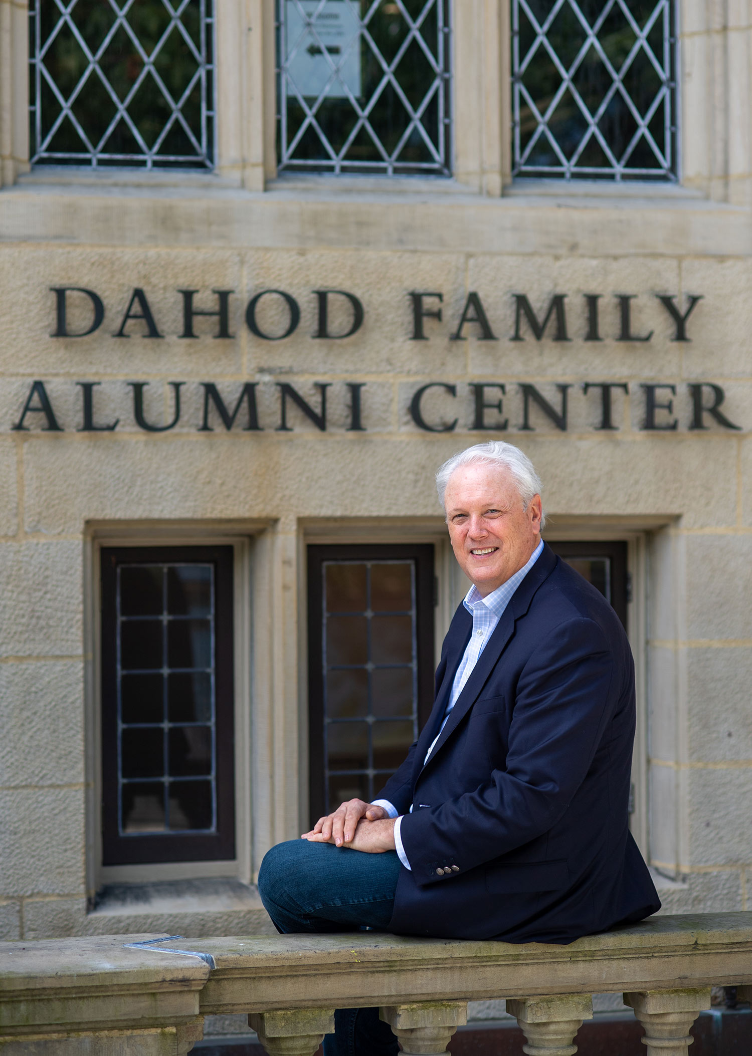 Steve Hall, VP in alumni relations who helped spearhead the hugely successful BU fund raising campaign, is retiring. Here he sits on one of the railings outside of the Dahod Family Alumni Center at the Castle on July 31, 2020. He wars a blue suits, and ornate windows are seen on the building behind hime with the sign "Dahod Family Alumni Center"