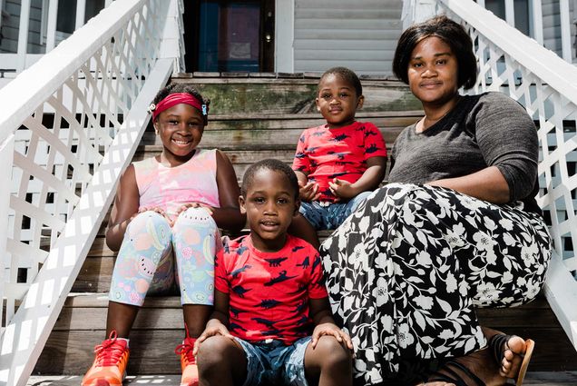 Abiba Braimah, a patient who took aspirin when pregnant with her two boys, both of whom were born near or full-term. She also has a daughter who was born prematurely, weighing only four pounds, before Braimah found the BMC doctor who prescribed aspirin. Braimah is pictured here with her three children, sitting on steps outside of her home. Her young boys wear matching red shirts with a dinosaur print, and her daughter smiles in a red head band and pink shirt.