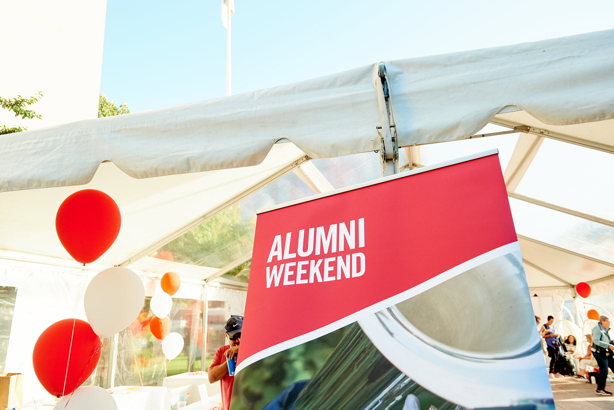 Photo of a read and white "Alumni Weekend" sign from 2019. Sign sits at the entrance of a white check-in tent, balloons and people checking in are seen in the background.