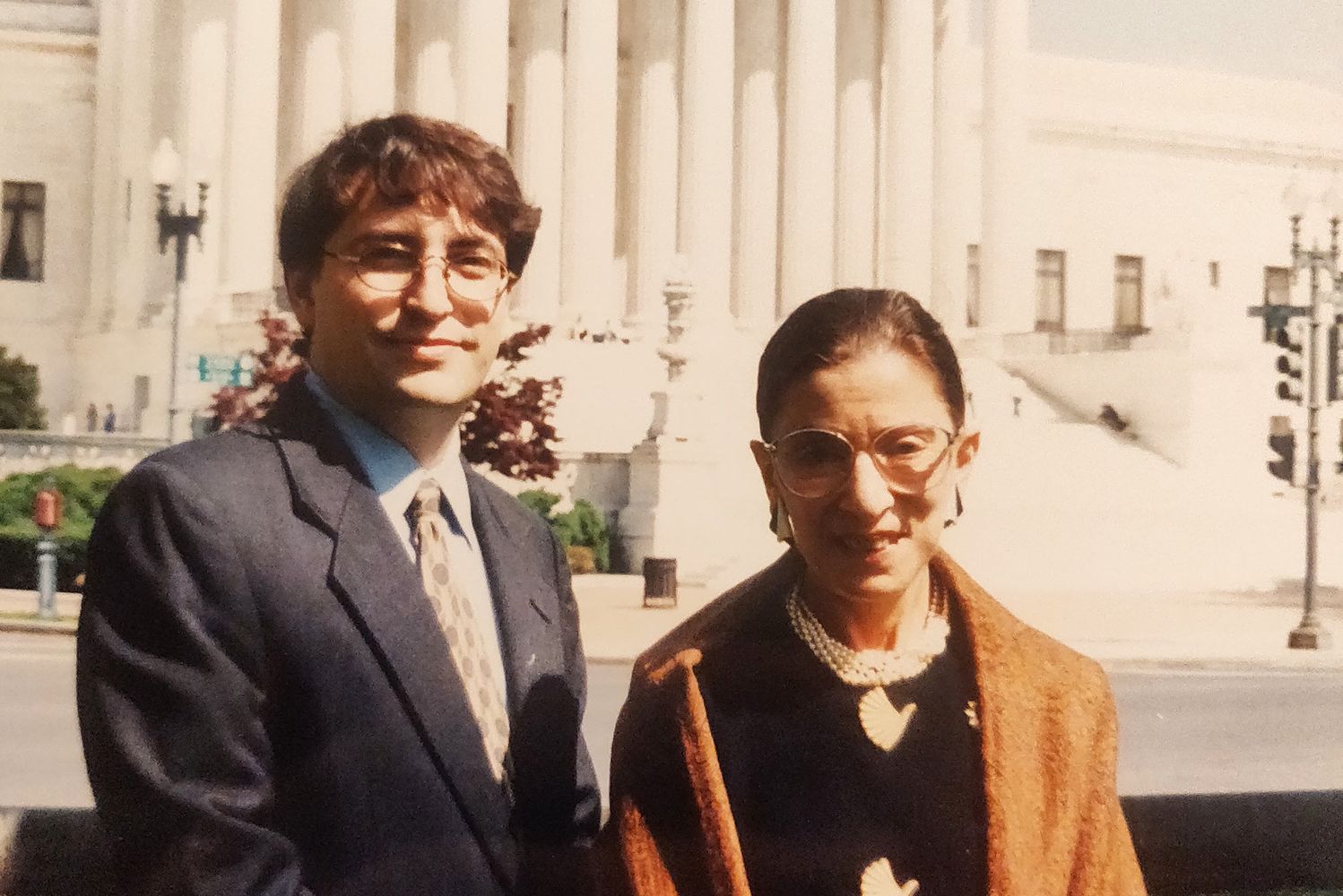 Photo of a printed photo of Jay Wexler (left) with Supreme Court Justice Ruth Bader Ginsburg in 1999, when he was one of her four clerks. Behind them is the facade of the Supreme Court. RBG wears a mustard colored shawl and Wexler wears a suit and tie.