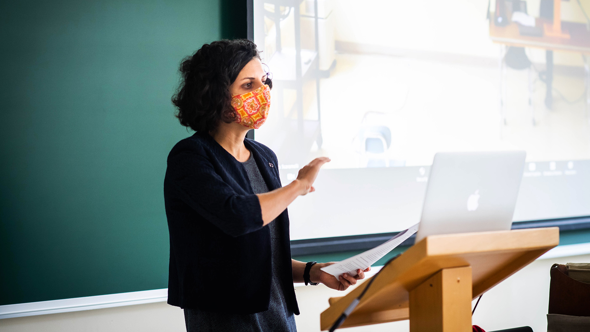 Photo of Caterina Scaramelli teaching an anthropology class, "Culture & Environment “ on September 22, 2020. She wears a bright orange mask as she lectures to her computer and the students in the class.