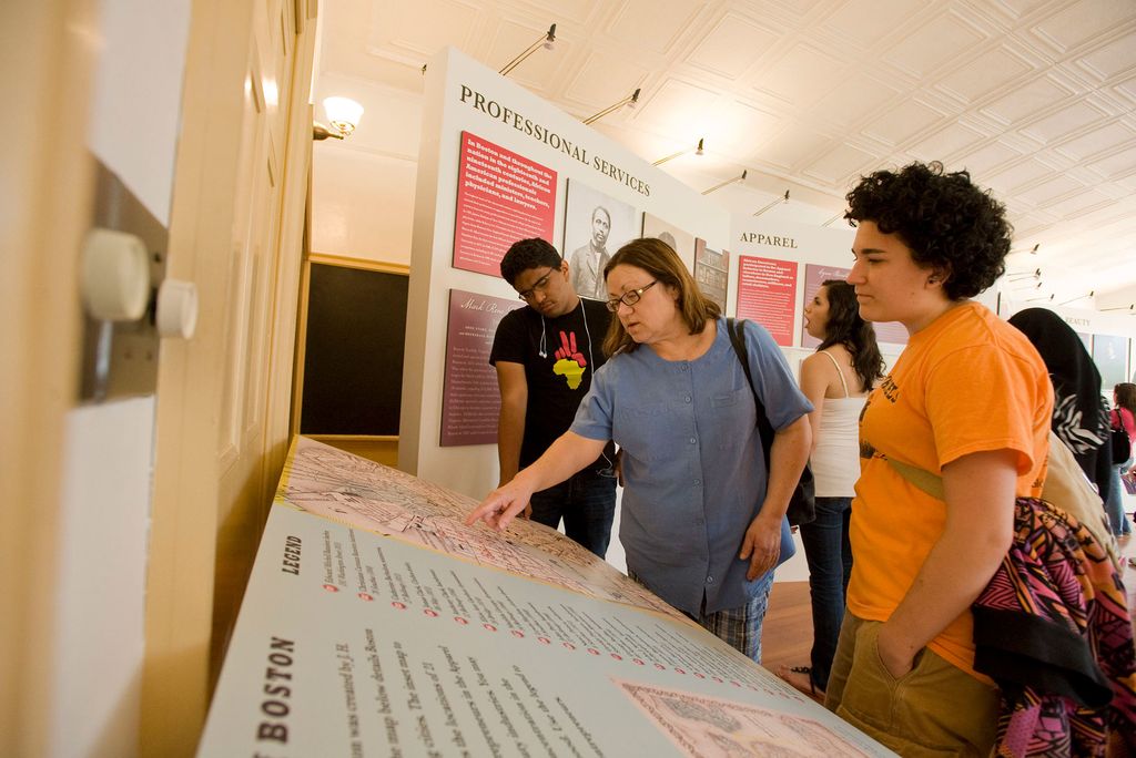 Photo of professor Mary Beaudry teaching MET students while on a walking tour of the Black Heritage Trail. She points at an exhibit description and wears a blue shirt.