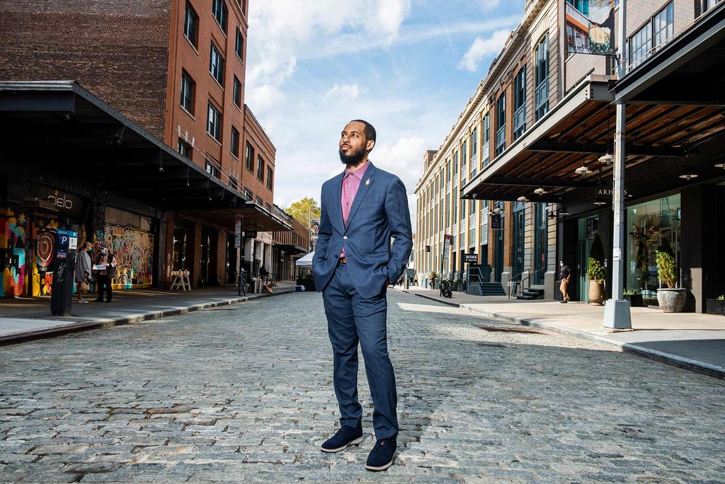 Photo of Jonathan Priester (COM’10) in blue blazer, pink button down, and gold lapel pin. He stands with his hands in his pockets, looks off to the middle distance, in a gray cobblestone street. The sky is blue behind him and some folks are seen on the sidewalk in the far distance.