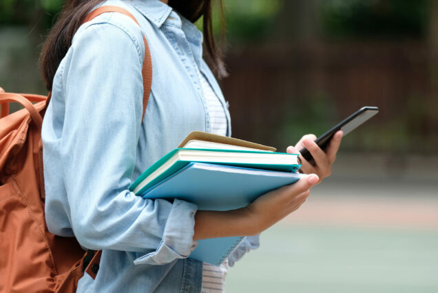 Photo of a student in a blue long sleeve shirt and wearing a backpack holding a stack of books.
