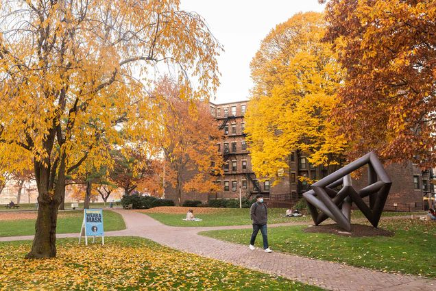 Photo of a student in a jacket and wearing a backpack walking down a path along BU beach near a sculpture and a sign that says "wear a mask." The trees are filled with bright orange and yellow leaves.