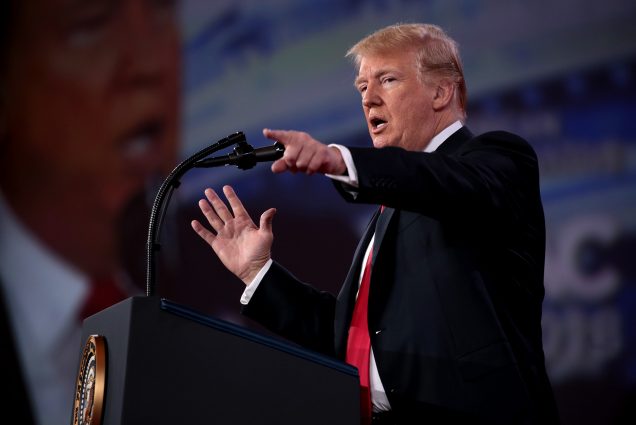 Photo of President of the United States Donald Trump speaking at the 2018 Conservative Political Action Conference (CPAC) in National Harbor, Maryland. He stands at a podium in front of a big screen and points at the crowd. He has a suit and red tie on.