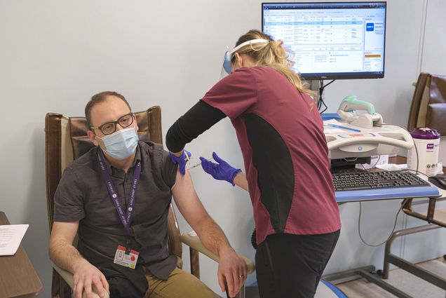 Photo of Joshua Barcoas of BMC getting a dose of the coronavirus vaccine. He sits in a chair with a blue face mask and his lanyard as a fellow healthcare worker gives him the shot.