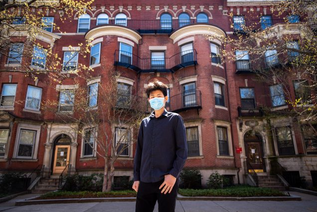 A photo of Jiaxin Tong wearing a mask standing outside of a row of brownstone residences
