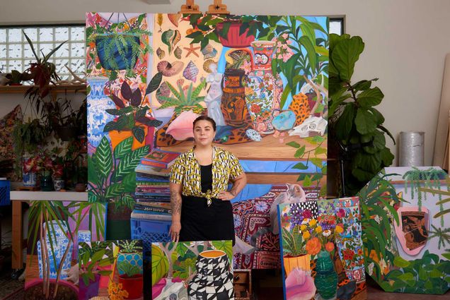 Photo of Anna Valdez in a cheeta print tied crop top and black pants standing amidst the plants and canvases of her studio. One huge canvas is seen behind her on an easel. Each canvas is filed with paintings of plants and statues, and bright color.