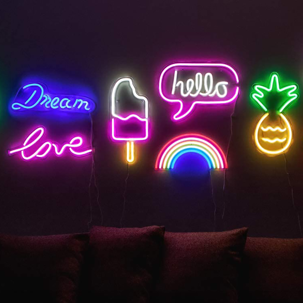A series of neon signs behind a couch. One says "Dream," another "Love" and "Hello" in a speech bubble. Others include a pink and yellow popsicle, a rainbow, and pineapple.