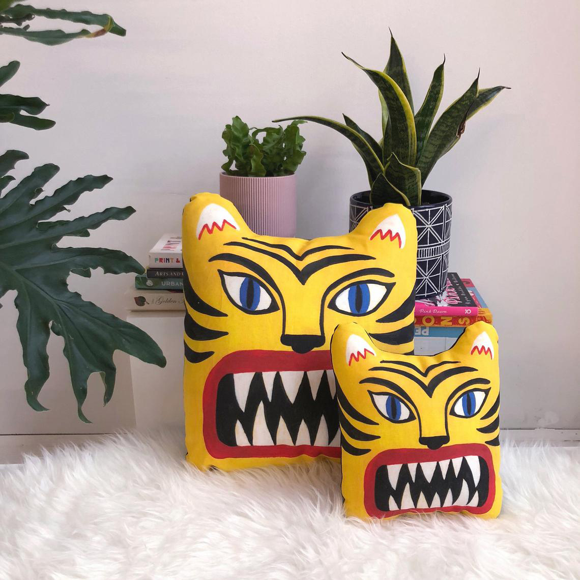 Photo of two lumbar support pillows that are shaped like tiger faces and have a bright orange tiger design on them. They rest on a white furry rug in front of some house plants.
