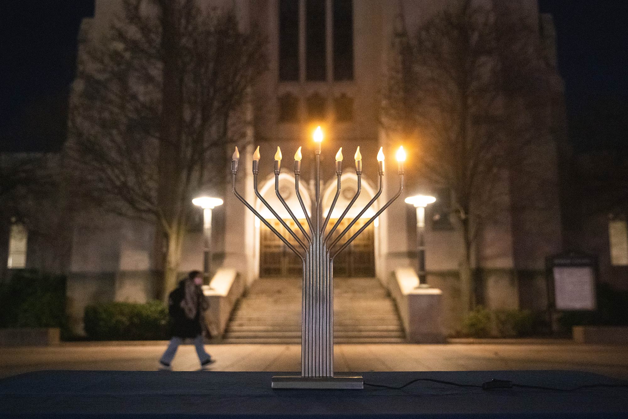 Photo of an electric menorah on a table in front of Marsh Chapel at night. The center and rightmost candle are light. A blurred person walks by in the background.
