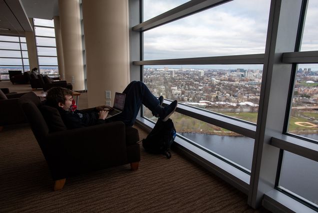 Photo of Ryan Blake (QST’21) working on a final project for the semester in the Stuvi2 Lounge. He wears a mask and has his feet on the window sill as he works on his laptop. A view of Cambridge across the Charles River is seen outside the window; it's partly cloudy.