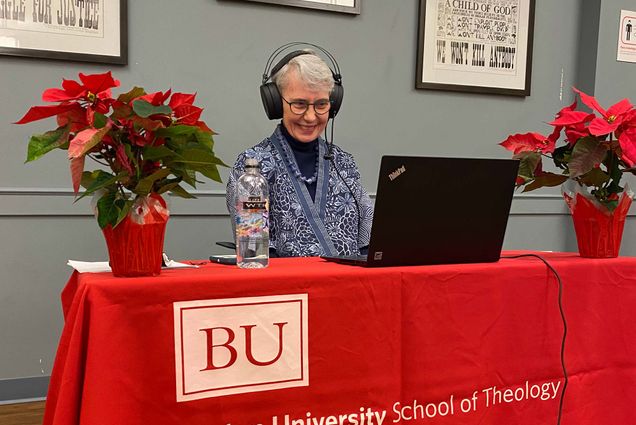 Photo of Mary Elizabeth Moore delivering her last lecture as dean before stepping down December 31. She delivered it virtually from the newly dedicated Mary Elizabeth and Allen Moore Community Center (lower level of STH building). She sits in front of a black laptop with large headphones on and smiles at the screen. The table has a red tablecloth that says "BU School of Theology." A poinsettia is seen to Moore's right. She wears a blue and white floral blouse.