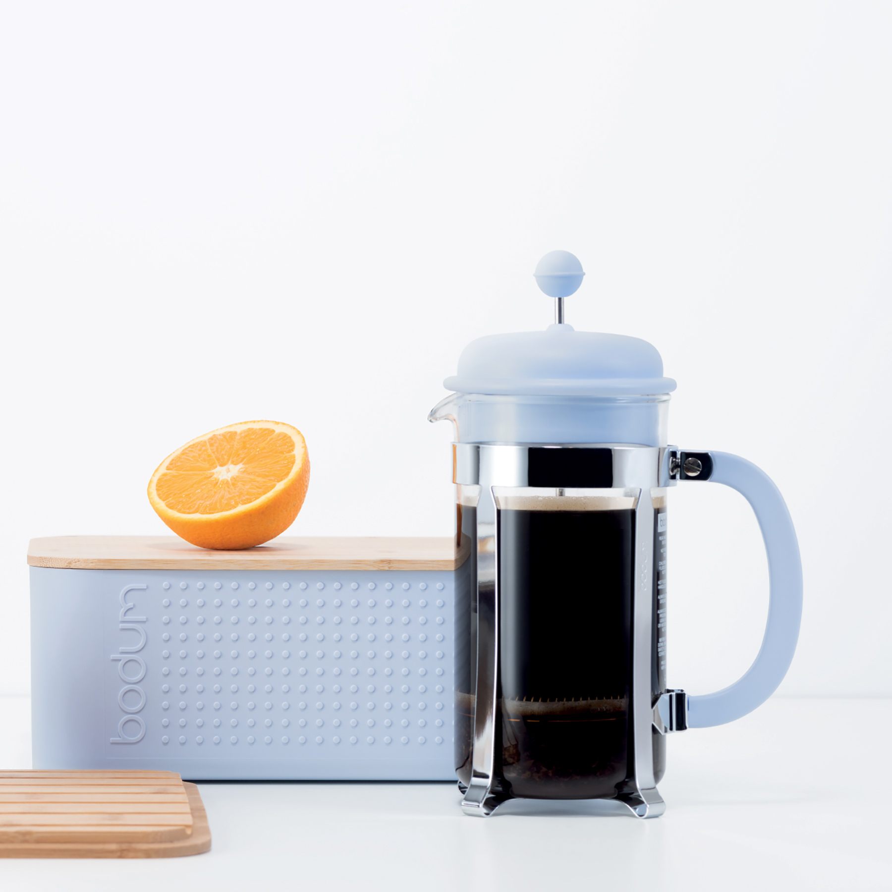 Photo of a light blue french press on a countertop with a clear coffee cup and orange.