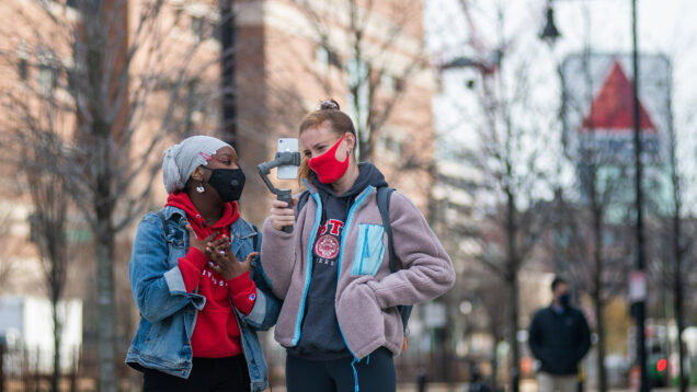Photo of Hikima Lukomwa (SAR’22), community linked coordinator, in a red sweatshirt and jean jacket, with Jillian White (COM’22), in a pink fleece, a community development leader, while they give a virtual tour use a handheld phone tripod. The Citgo sign is seen blurred in the background.