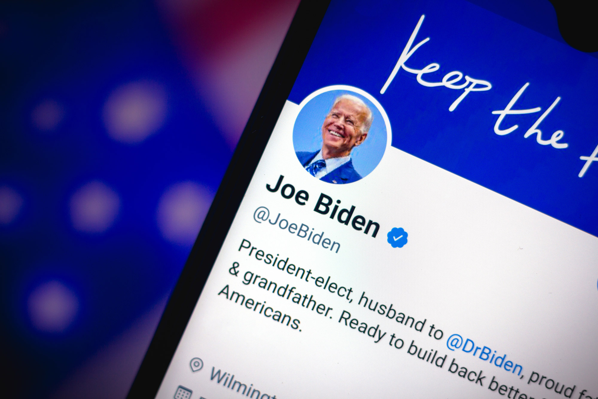 A photo of Joe Biden's twitter account displayed on the screen of a smart phone. The phone is held in front of an American flag.