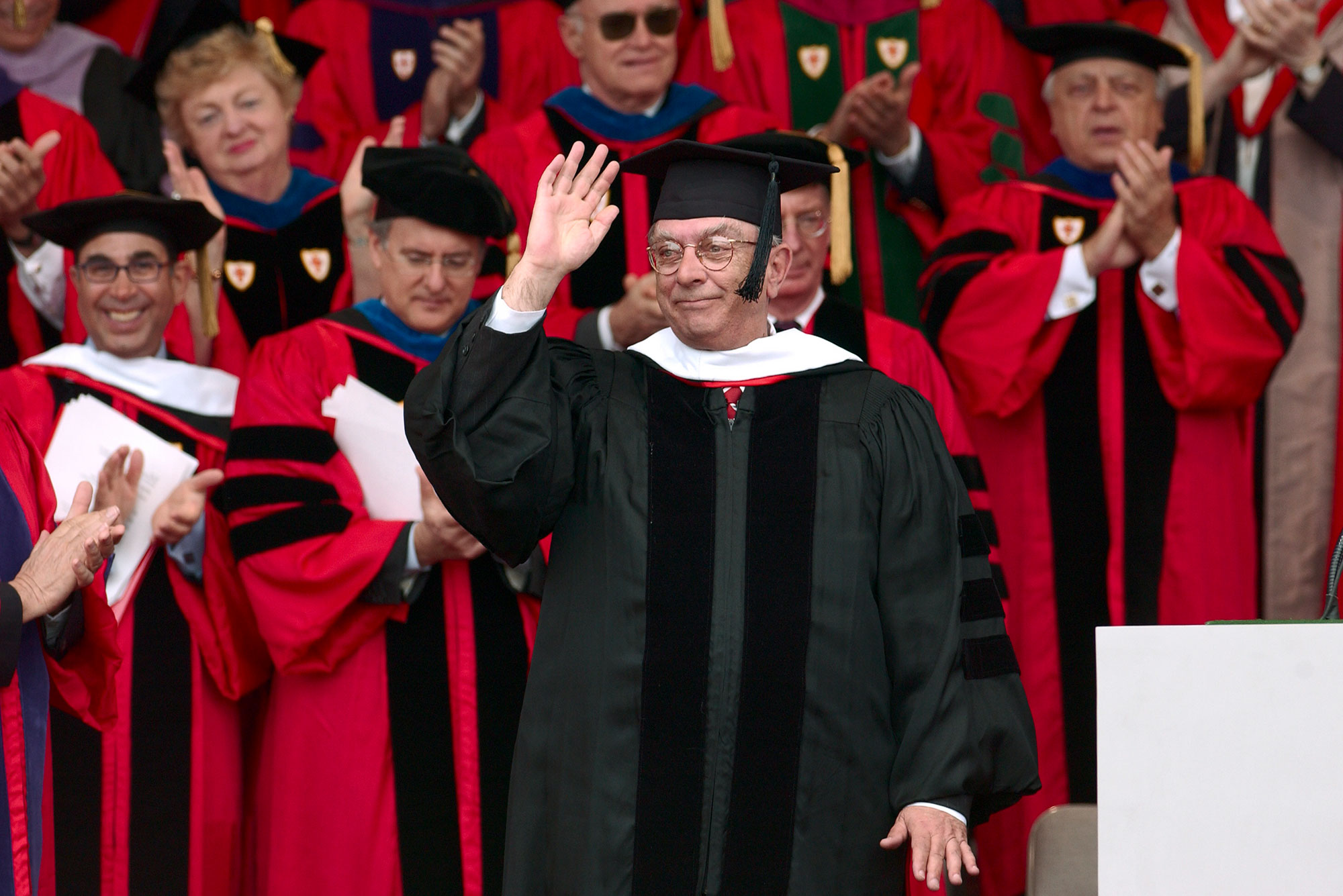 Photo of Jon Westling receiving an honorary Doctor of Humane Letters Degree in 2003. Westling wears a black robe and cap and waves with his right hand. A crowd of faculty in red regalia are seen behind him.