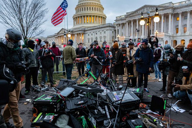 Photo of the rubble left in the aftermath of pro-Trump protestor attack on the press pool outside the Capitol building. Journalist stand around in a circle, at the center, a pile of their destroyed TV production gear sits on the ground.
