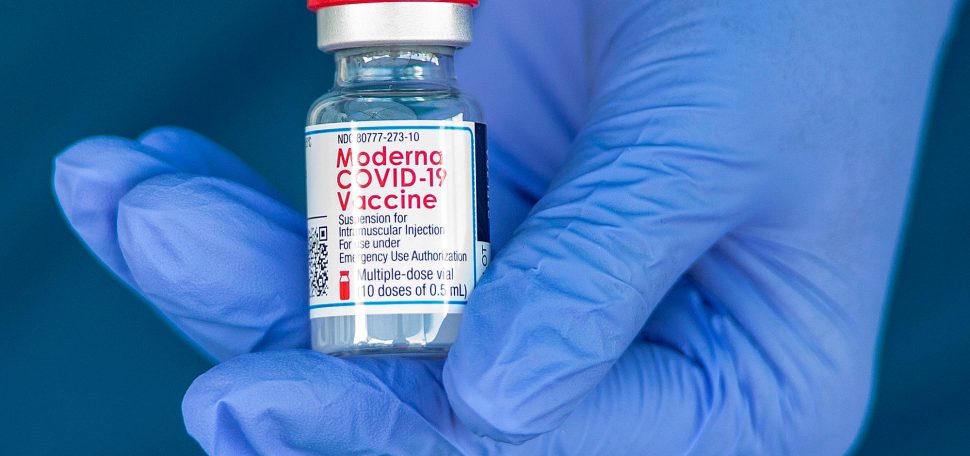 Photo of the gloved hand of a registered nurse holding a vile of the Moderna COVID-19 Vaccine before giving the vaccine to healthcare workers in Anaheim, Calif. on Friday, January 8, 2021. The small glass vile has a red and silver top; the label reads “Moderna COVID-19 Vaccine: suspension for intramuscular injection, for us under emergency authorization. Multiple doses vial (10 doses of .5 ml)”