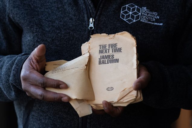 Photo of Dean Elmore holding a tattered copy of "The Fire Next Time by James Baldwin. The photo is a tight shot on Elmore's hands as he opens to the book's yellowed title page.