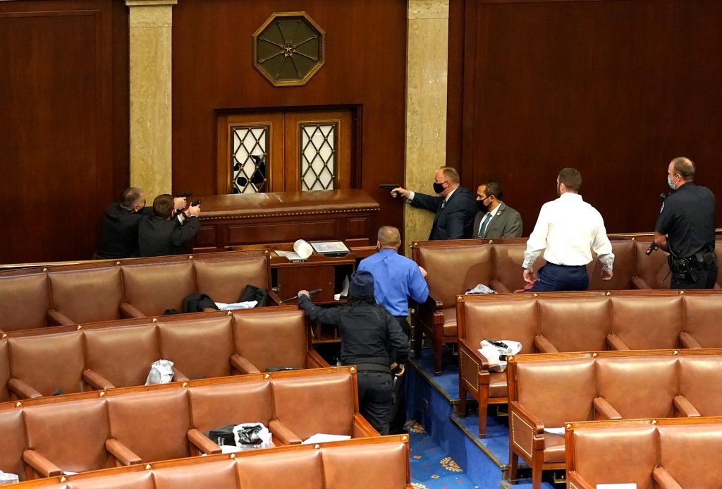 Photo of U.S. Capitol police officers pointing their guns at a door that was vandalized in the House Chamber during a joint session of Congress on January 06, 2021 in Washington, DC. A group of men and women rush towards the vandalized area to protect the chamber. The chamber seats are empty.