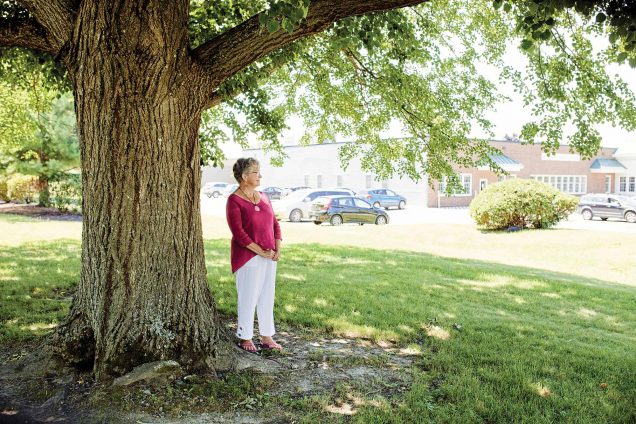 Photo of Bonnie-Jean Brooks in a pink blouse and white pants standing under a large tree looking off into the the distance.