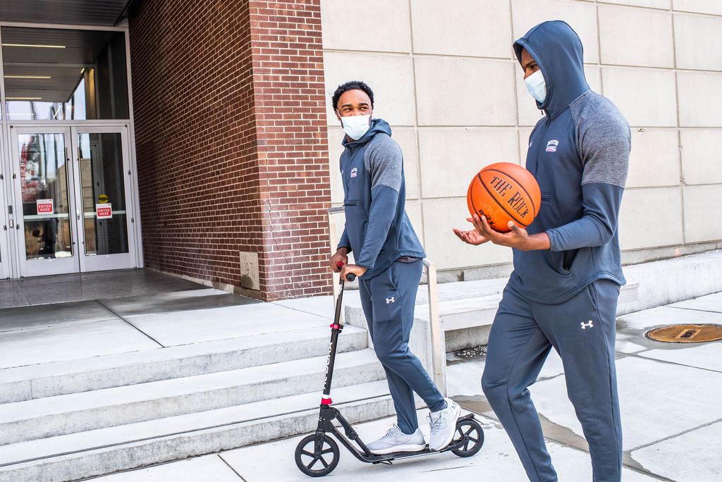 A photo of Walter Whyte and Jonas Harper heading to practice together. One is riding a scooter, the other is holding a basketball. Both are wearing masks.