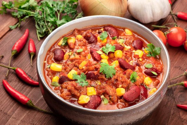 Detail photo of a bowl of Chili Con Carne on a wooden table with recipe ingredients.