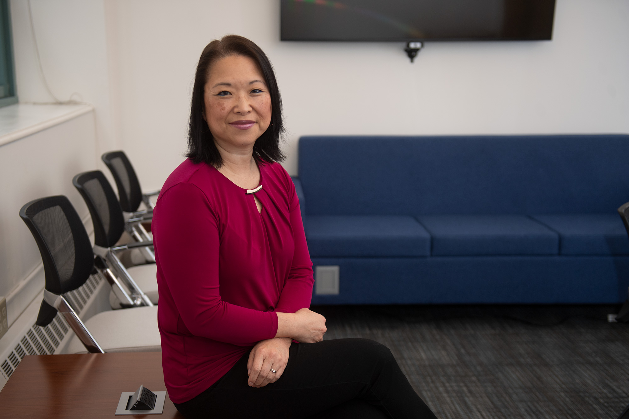 Photo of Maria Erb, in a scarlet blouse, smiling as she sits on a side table. In the background, a blue couch is seen.