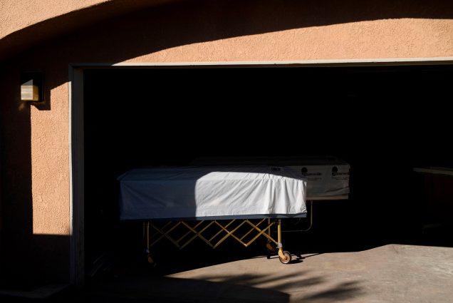 Photo of a casket covered with a white sheet over it on a dolly at the edge of a garage; it is partly in the shadows and the garage is darkened.