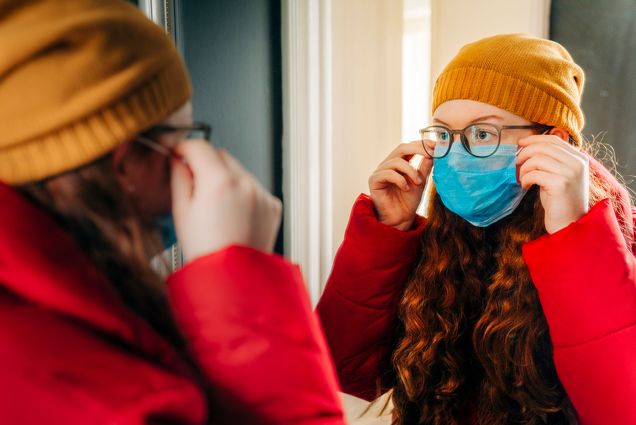 Photo of a young women with long, wavy, red hair looking into the mirror as she puts on a blue face mask. She wears a mustard yellow beanie and thick jacket, and appears to be getting ready to go outside in the cold.