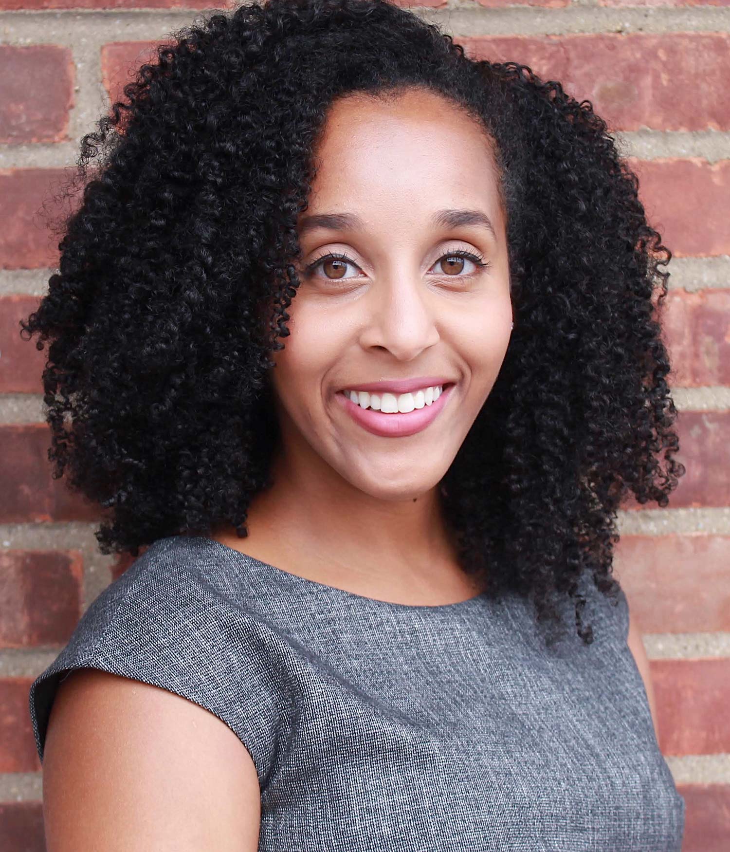 Headshot of Alexis Redman (Questrom’14), head of the BU Young Alumni Council, in a gray textured blouse, smiling as she stands in front of a brick wall.