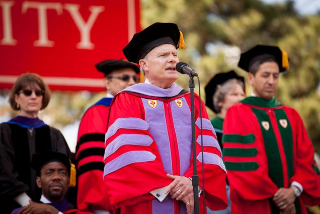 Photo of Goldman School of Dental Medicine Dean Jeffrey Hutter presenting candidates during the 2015 Boston University Commencement at Nickerson Field on May 17, 2015. Hutter wears BU regalia, a red rob with purple stripes on the sleeve and down the front, and a black octagonal hat with a gold tassel. He stands at the mic as he speaks, other faculty are seen blurred behind him.