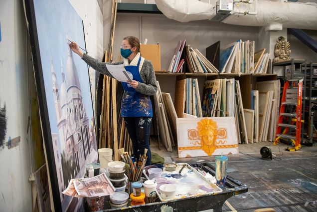 Photo of Trish Hawkins (CFA’21), wears a gray and white sweater and blue painting smock, as she work on her painting of the Santa Maria della Salute, a Roman Catholic church in the city of Venice, Italy, which has a large central white dome. Hawkins works in the Main Print Shop in Booth Theater February 10, behind her stacks of canvases are seen stored; one painting shows an orange knight from a wall frieze.