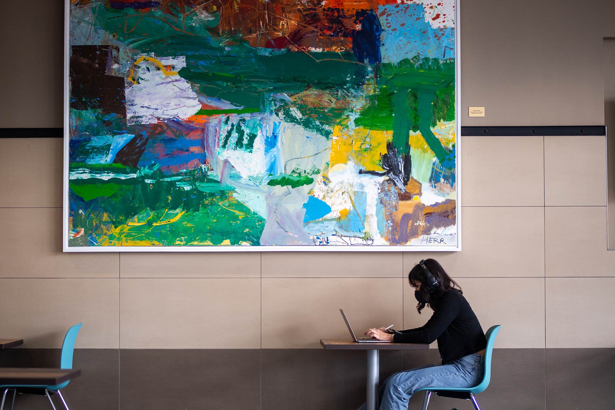 Photo of a student in a black shirt in jeans sitting alone at a table with a face mask on as they attend virtual class. A large abstract painting hangs on the wall behind them.