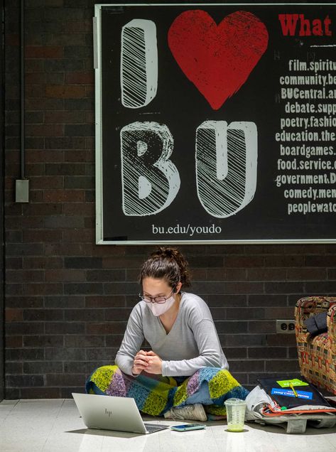 Photo of a student sitting criss-crossed on the ground as she attends a virtual meeting on her laptop. Beside her, a plastic cup and a stack of books and papers on top of her backpack is seen. Behind her, a big black sign reads “I Heart BU” with the url bu.edu/youdo.