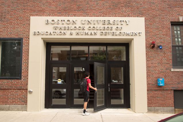 Photo of a student in a red shirt and black shorts wearing a black backpack opening the door to Boston University Wheelock College of Education & Human Development at 2 Silber Way.