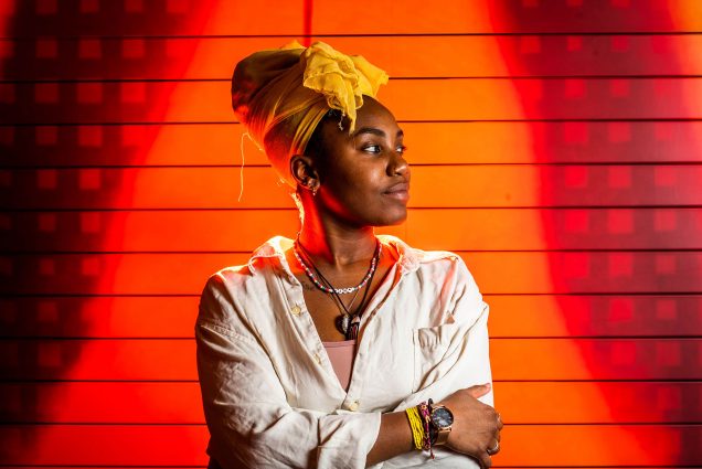 Photo of Curator-in-residence of the Jar Ireon Roach (CFA’21) with her arms folded across her looking off to her left; she is brightly lit by orange lights and wears a yellowy hair wrap.