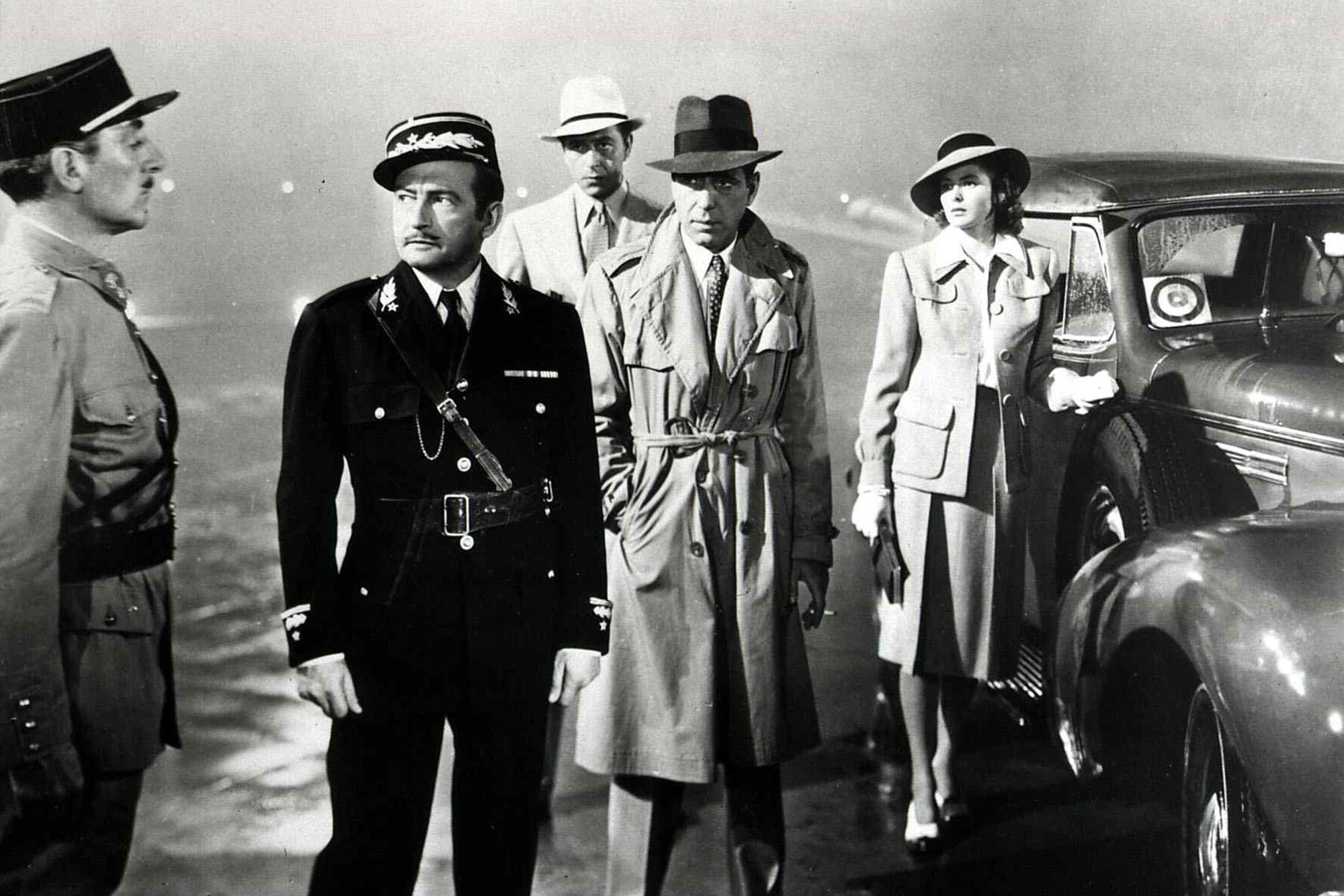 Black and white still from the film “Casablanca.” In the culminating scene, Jean del Val, Claude Rains, Paul Henreid, Humphrey Bogart, and Ingrid Bergman are seen. The men wear trench coats and hats, and the officers wear uniforms.