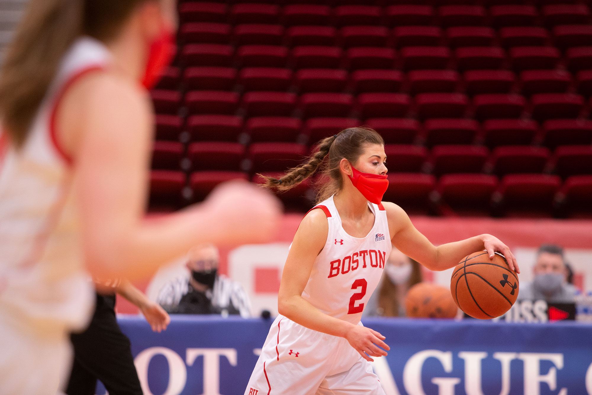 Photo of Katie Nelson dribbling the ball down the court during the game against Lehigh; she wears a red face mask and a jersey with the number 2. Another BU player is seen blurred in the left foreground.