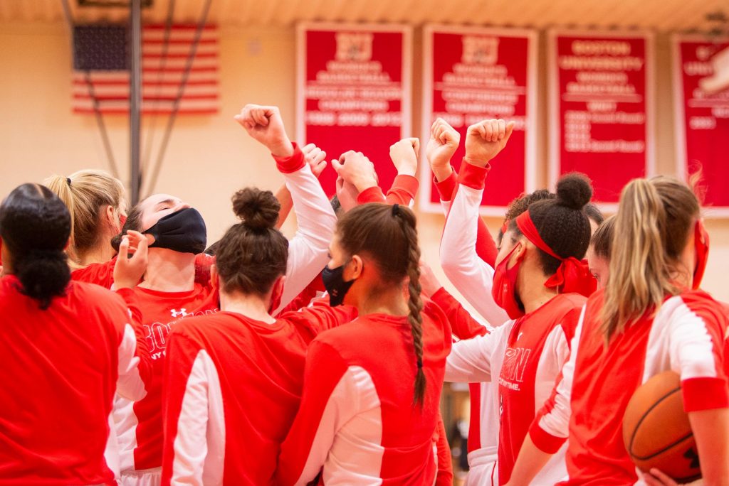 Photo of a team huddle of BU Women’s basketball players before the patriot league championship game. They wear warm-up gear and red face masks.
