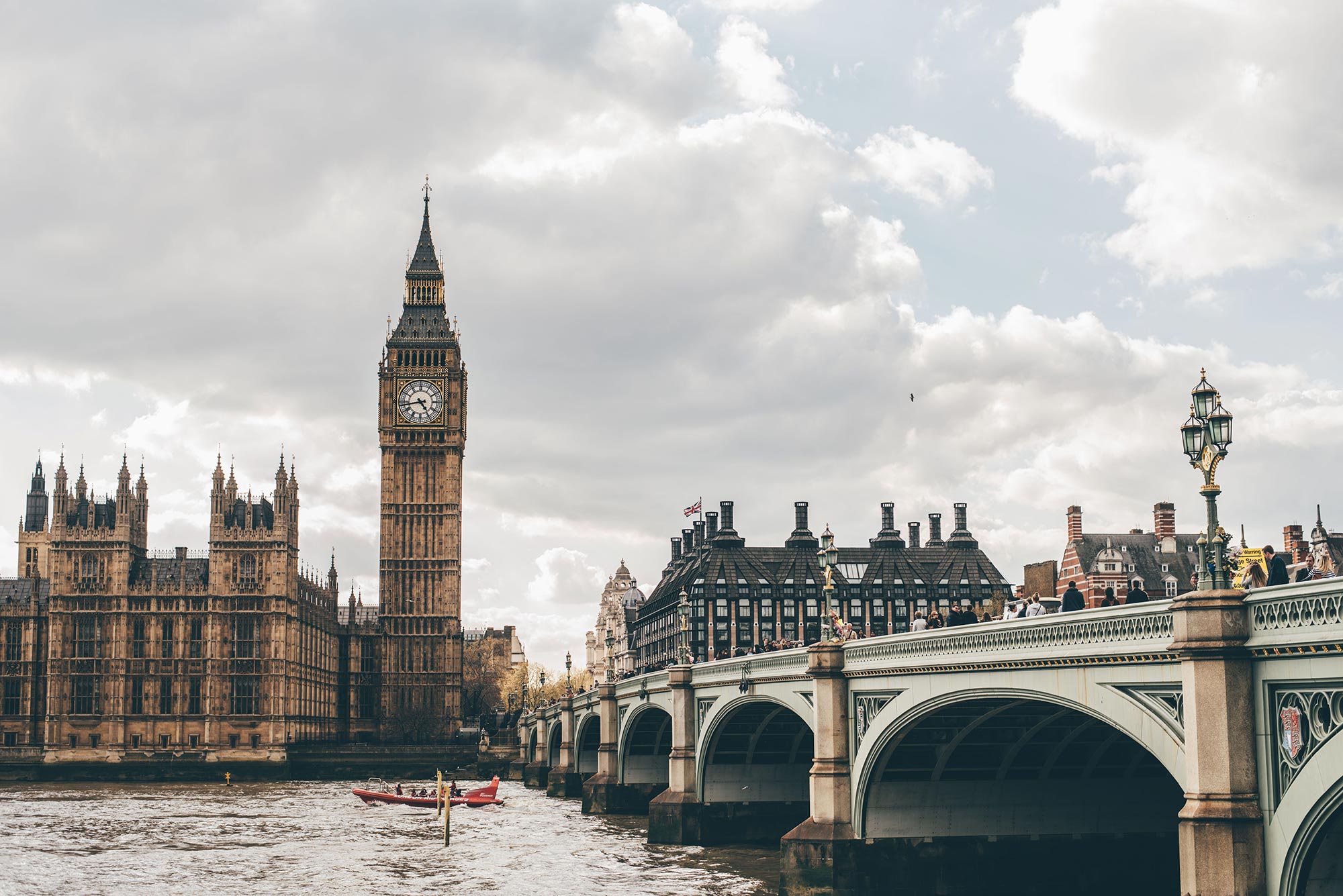 Photo taken from across the Thames River of Big Ben with an ornate bridge on the right; the heads of people walking over the bridge are seen. In the river, a small red boat with people moves to the left; the sky is grayish and cloudy.