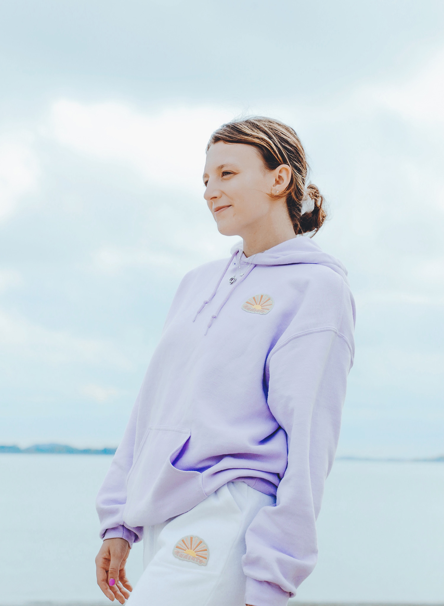 Photo of Kate Silvestri (Questrom’23) modeling the light purple Lotus hoodie, one of the pieces in her Radiate clothing line. Behind her, a gray sky and body of water are seen.