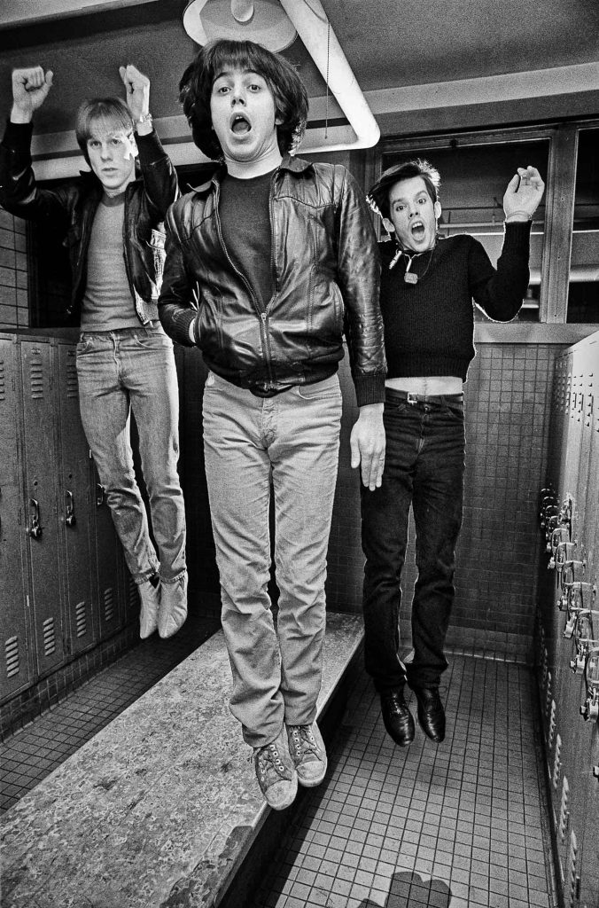 Black and white photo of the Boston band the Neighborhoods. Three young men dressed in black and leather jackets jump in the air in what looks like a locker room.