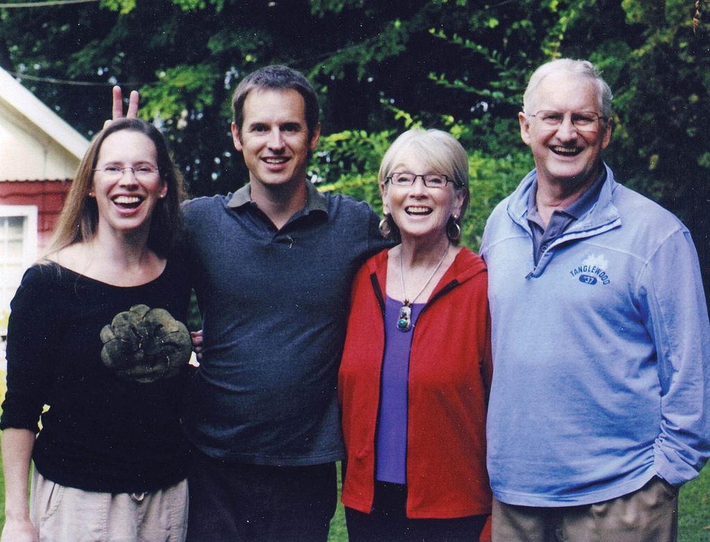 A somewhat faded photo of the Douglass family standing outside with trees behind them. The brother makes bunny ears behind his sister, and the two parents stand smiling at the right. A corner of their red Stockbridge, Mass., guest house is seen in the background on the left.